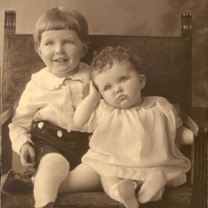 Two children sit on a chair facing the camera. The boy on the left is bigger and older than the girl on the right. The boy is smiling; the young girl rests her head on her hand.